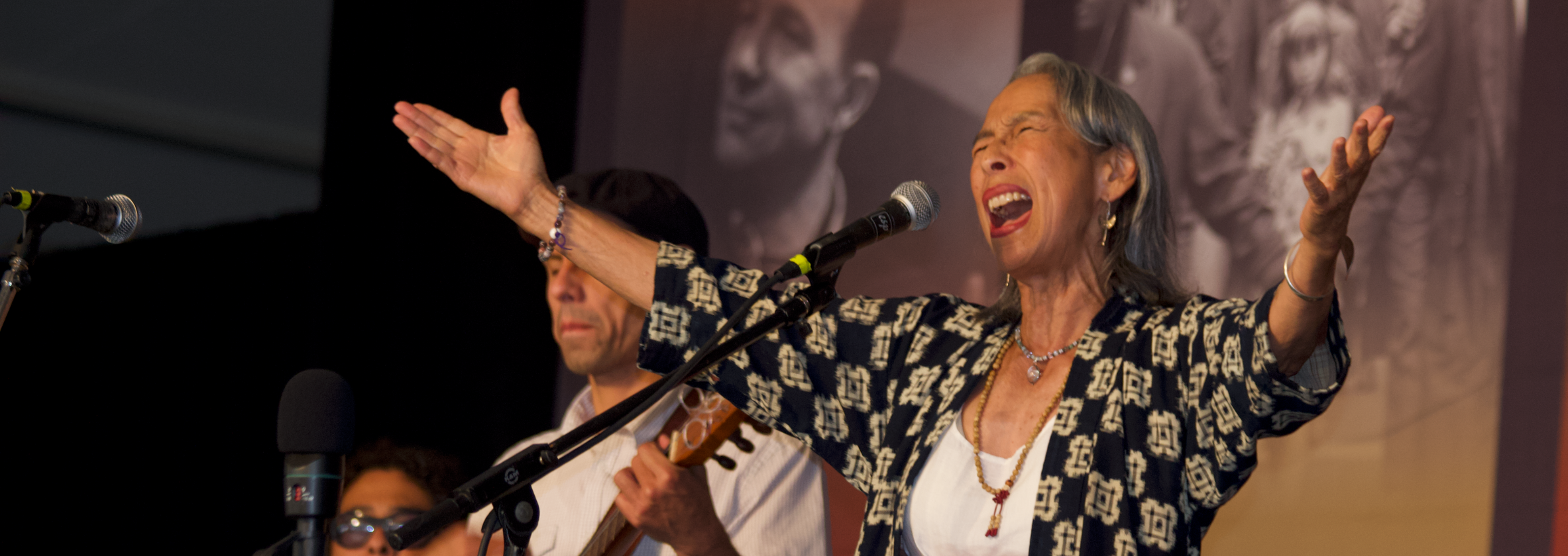 nobuko miyamoto singing on stage with band behind her. Her arms are spread out open