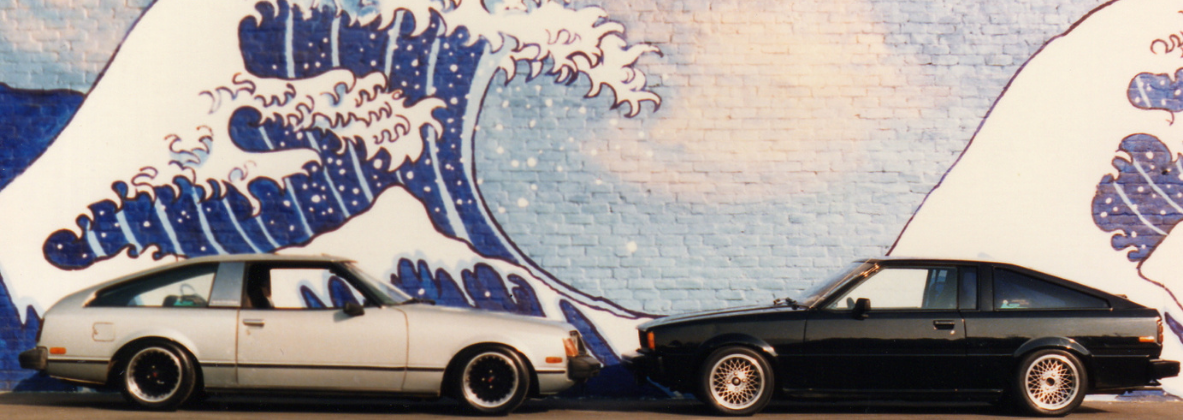 Two vintage cars are parked nose to nose in front of a mural of a Japanese style ocean wave