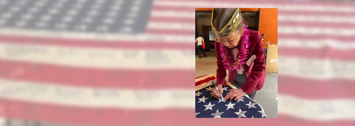 A veteran leans over a flag to sign it. This image is placed over a faded image of a flag with signatures.