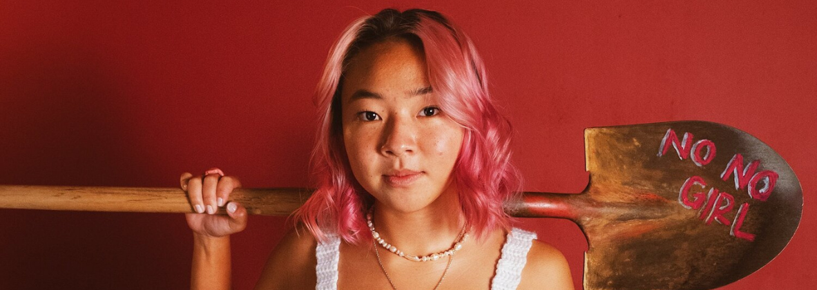 A girl with pink hair, holding a shovel over her shoulder with the words "No No Girl" painted on it.