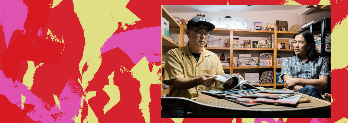 An image is framed by a colorful red, pink, and yellow background. In the image, two people sit at a table covered in Giant Robot magazines. The person on the left is flipping through a magazine.