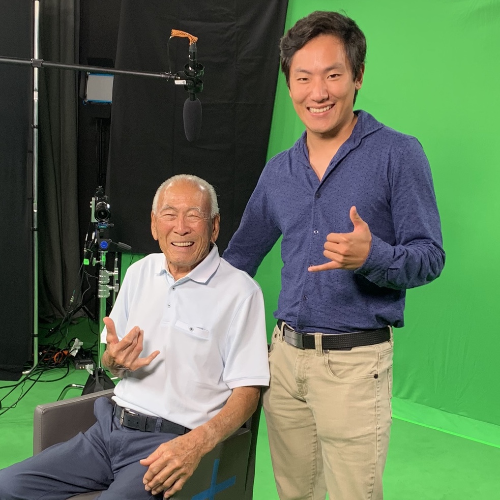 photo of Cole Kawana standing behind Lawson Sakai (seated in a chair), both are in front of a green screen with cameras in the background