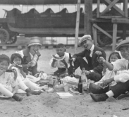 A group of early 1900s Japanese Americans sit around a picnic of glass bottles. They are holding watermelon and chopsticks.