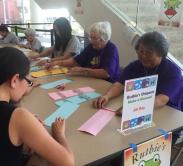 People sit around a table folding origami. Signs say "Ruthie's Origami."