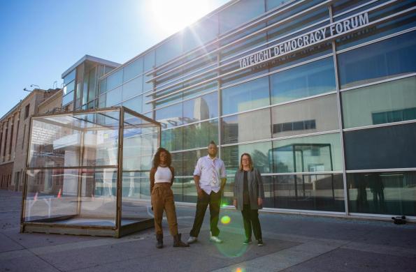 three people stand next to large glass box sculpture with writing engraved on the sides. NCPD building in the background