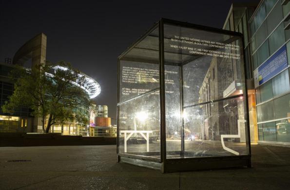 photo of glass box at night, box has written engravings on the side. JANM building and NCPD are in the background, box is in the foreground