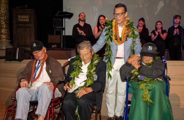 David Ono stands behind 3 veterans from the 442nd Infantry Battalion. They are each sitting in wheelchairs and they all are wearing leis around their necks, including David Ono.