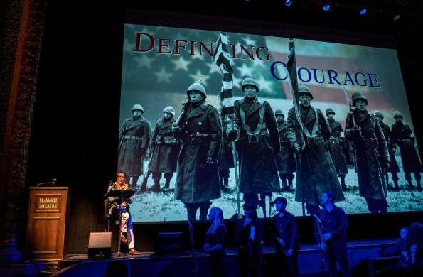 David Ono stands at a podium speaking to an audience. Behind him is a large screen projection showing an image of the 442nd Infantry Battalion and the title says, "Defining Courage"