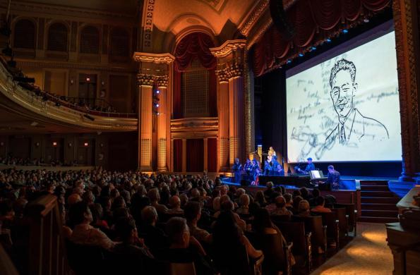 Large theater and audience sitting watching an orchestra. There is a screen on stage that shows an illustration of Stanley Hayami