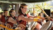 Smiling taiko drummers in red and yellow outfits stand at the ready in front of angled drums
