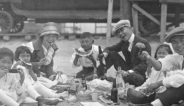 A group of early 1900s Japanese Americans sit around a picnic of glass bottles. They are holding watermelon and chopsticks.