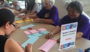People sit around a table folding origami. Signs say "Ruthie's Origami."