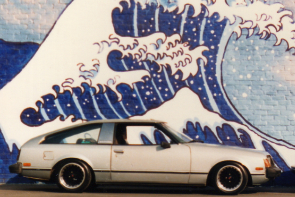 Two vintage cars are parked nose to nose in front of a mural of a Japanese style ocean wave