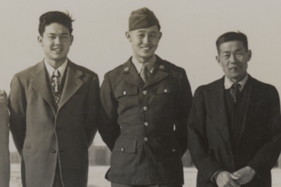 A black and white photo of a family of four Japanese Americans. One is in 442nd uniform of WWII. There are barracks behind.