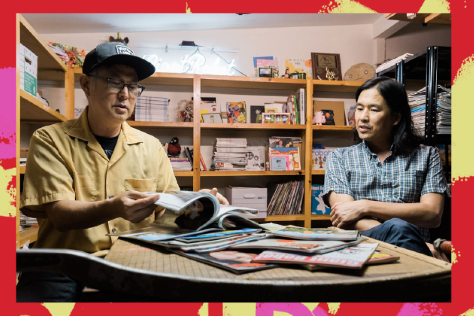 An image is framed by a colorful red, pink, and yellow background. In the image, two people sit at a table covered in Giant Robot magazines. The person on the left is flipping through a magazine.