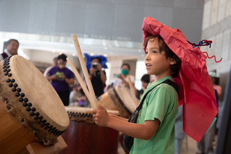 little boy playing taiko drum wearing hand made paper hat during natsumatsuri family festival