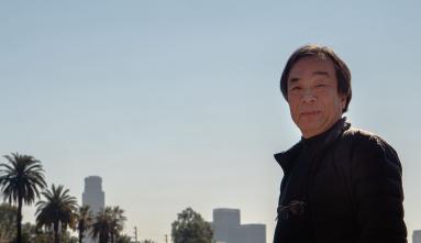 Photo of artist Masaki Fujihata in front of lake with palm trees and city skyline in the background