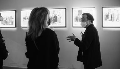 A black and white photo of BeHere / 1942 artist Masaki Fujihata giving a tour in front of illuminated images.