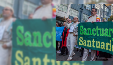 People of different faiths marching down a street, holding a banner that reads "Sanctuary" and "Santuario".