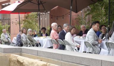 men and women have tea sitting at a long table listening to the speaker